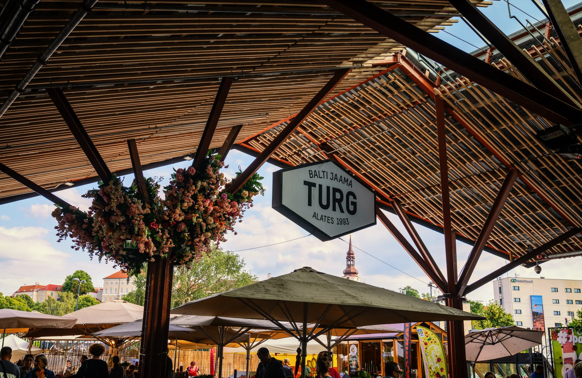 The wooden-canopied Balti Jaama Turg market hall in Tallinn with its sign, hanging flowers and market umbrellas, the railway station behind