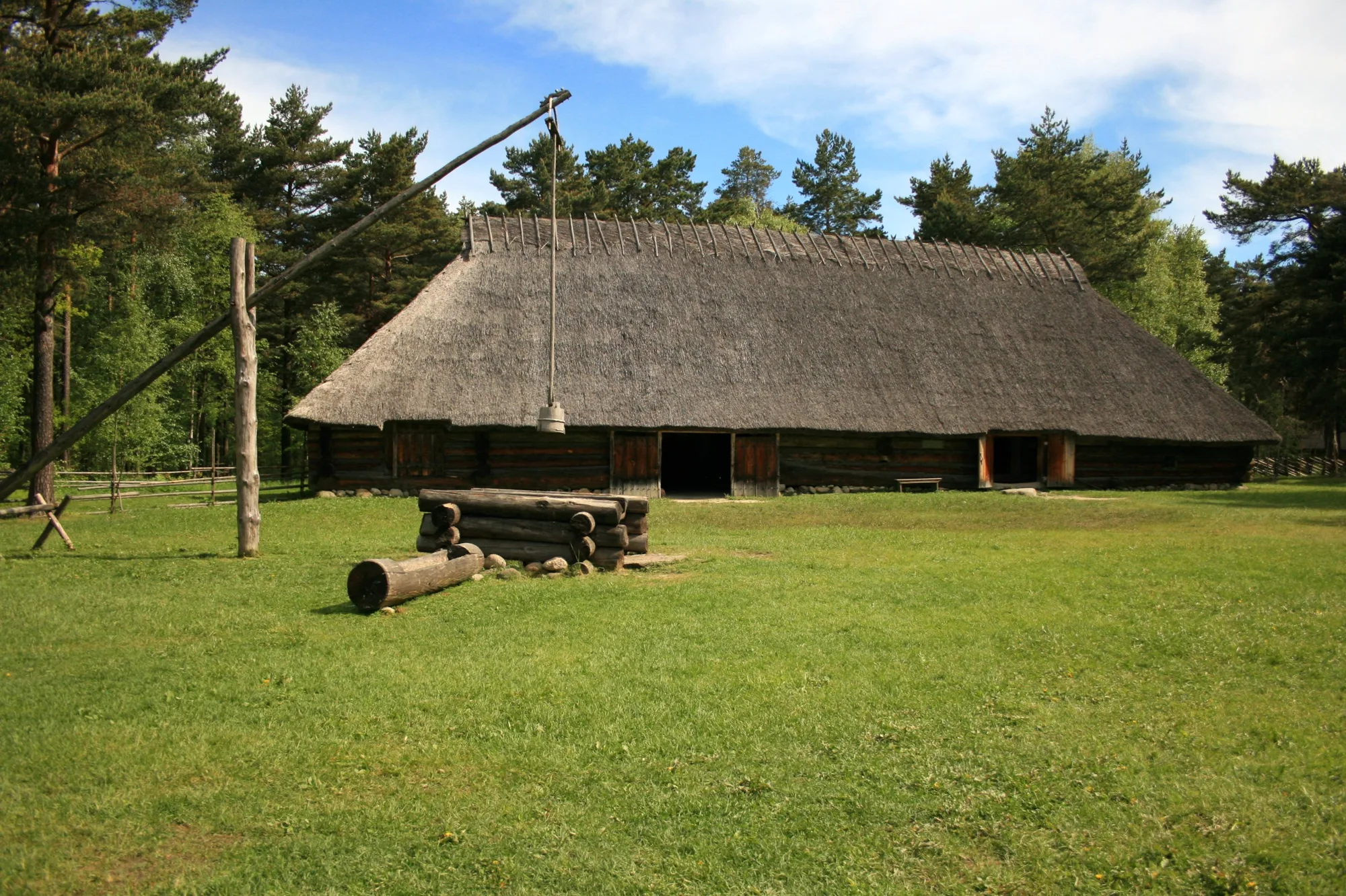 A historic thatched-roof log farmhouse with a traditional well sweep on a green meadow at the Estonian Open Air Museum, Rocca al Mare, Tallinn