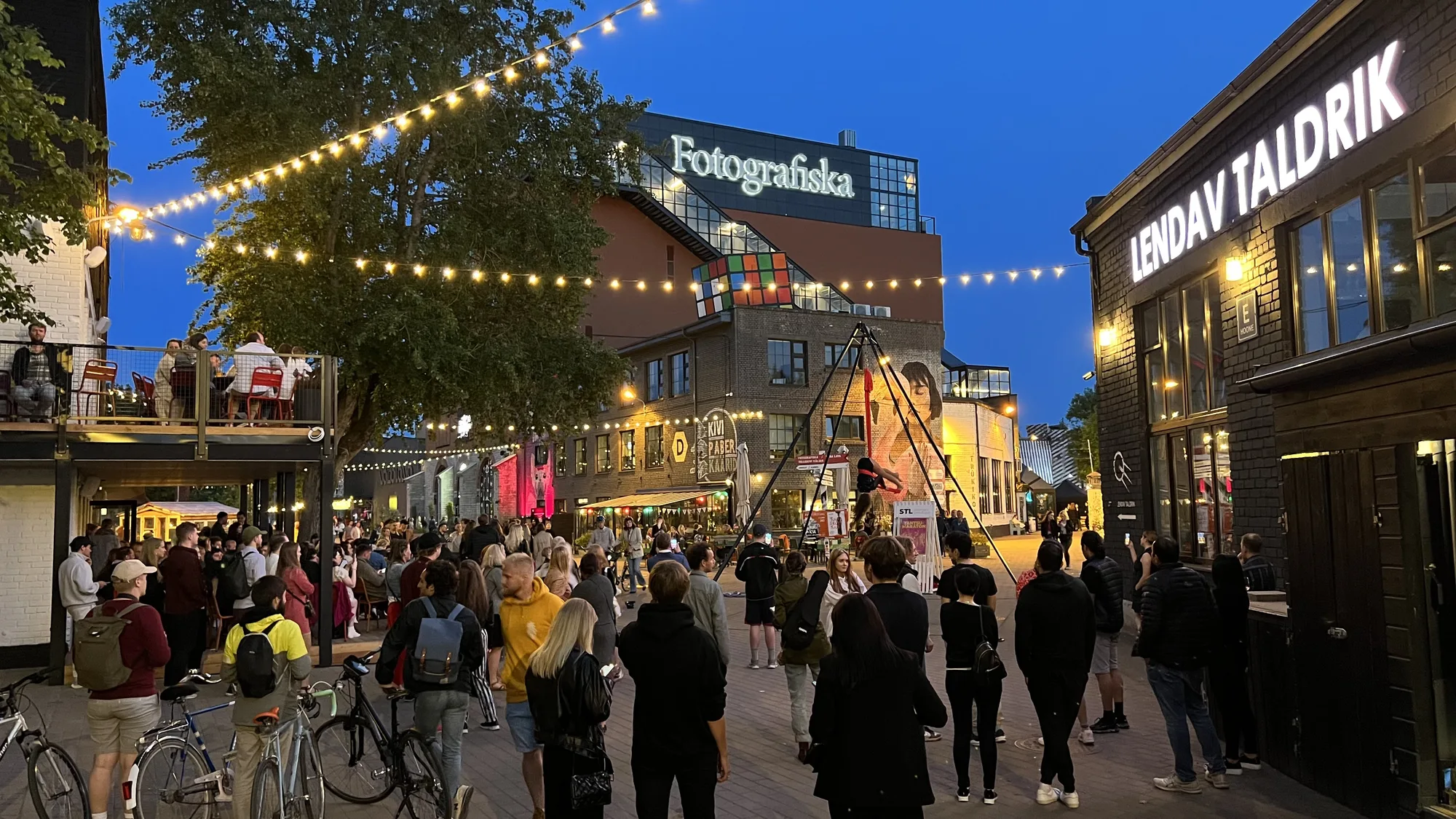 Crowds under string lights at dusk in Telliskivi Creative City, Tallinn, with the Fotografiska building and converted industrial buildings